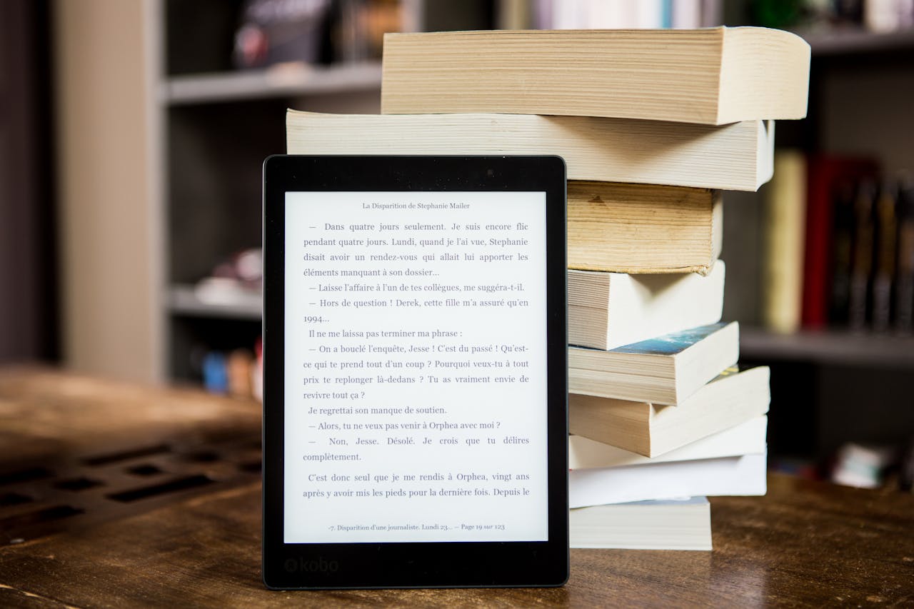 hero-img E-reader with text displayed next to a stack of physical books on a wooden table.